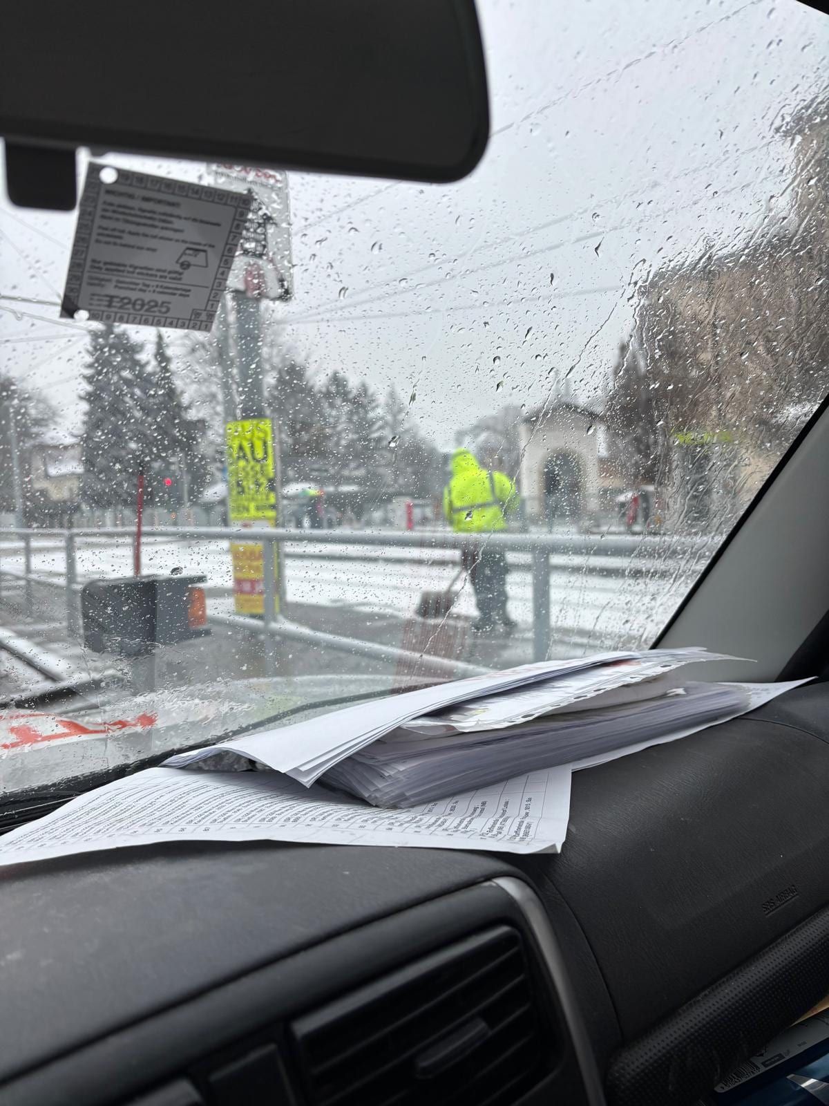 Blick durch die nasse Windschutzscheibe auf einen Mitarbeiter in Warnjacke, der mit einer Schneeschaufel einen Gehsteig räumt.