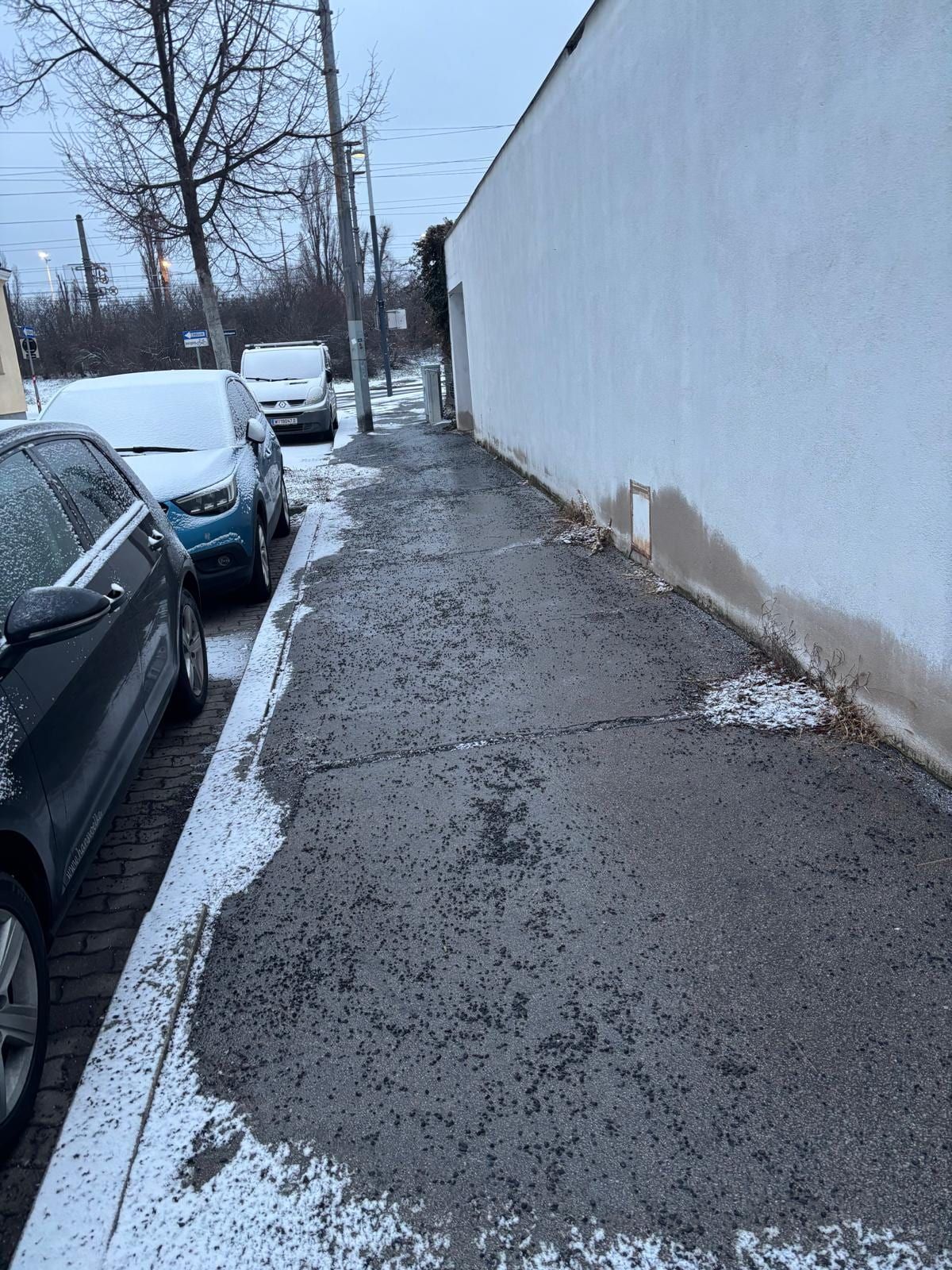 Gestreuter Gehsteig entlang einer weißen Mauer in der Dämmerung, links beschneite Autos.