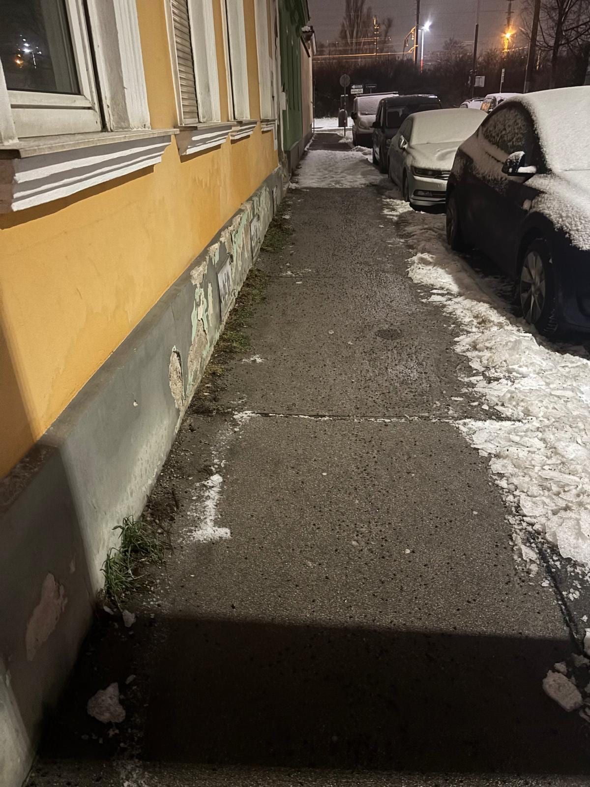 Geräumter und gestreuter Gehsteig entlang eines gelben Altbaus bei Nacht, daneben verschneite Autos und zur Seite geschobener Schnee.