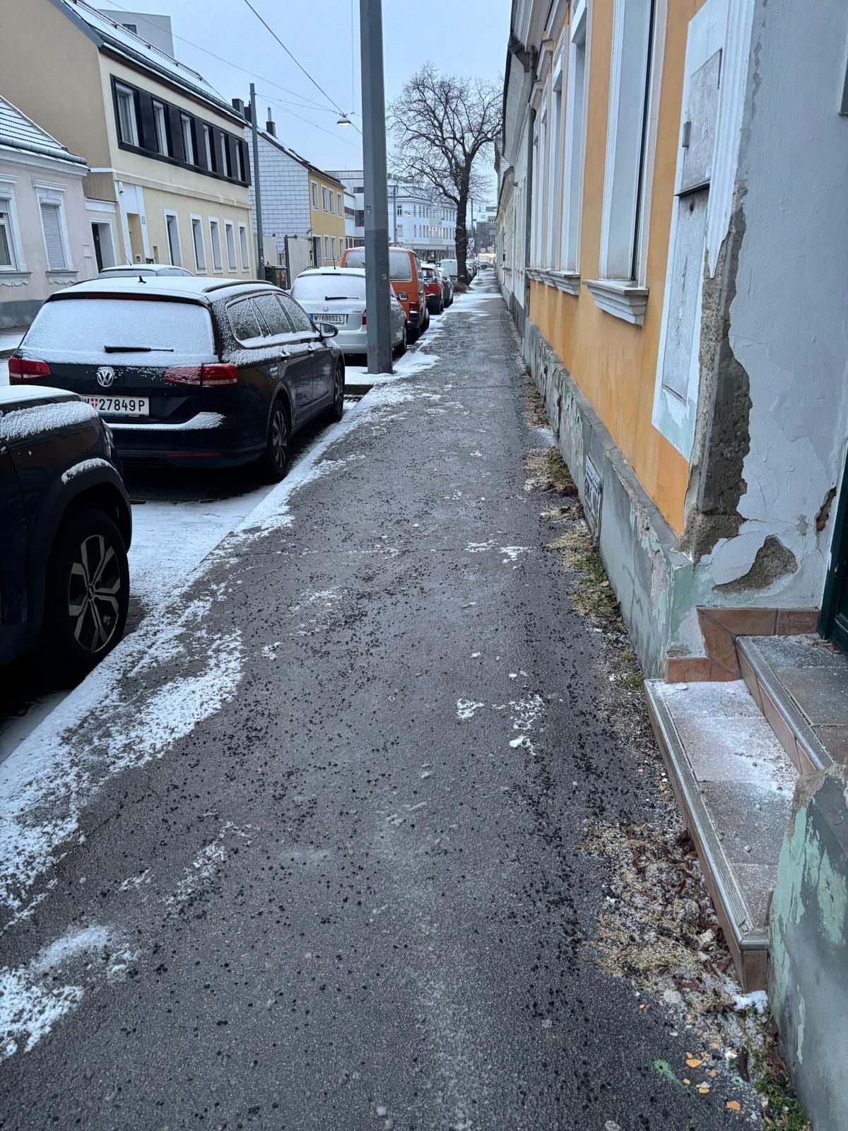 Geräumter und mit Split gestreuter Gehsteig entlang eines gelben Altbaus, daneben beschneite parkende Autos.