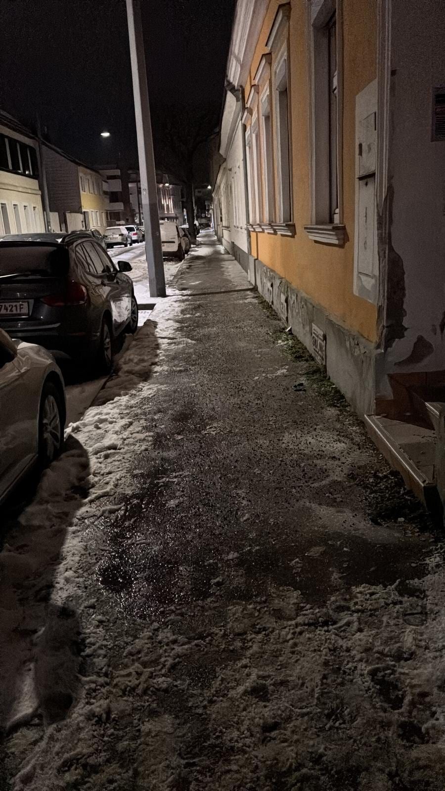 Gestreuter Gehsteig entlang von Altbauten bei Nacht, links parkende Autos unter Straßenlaternen.