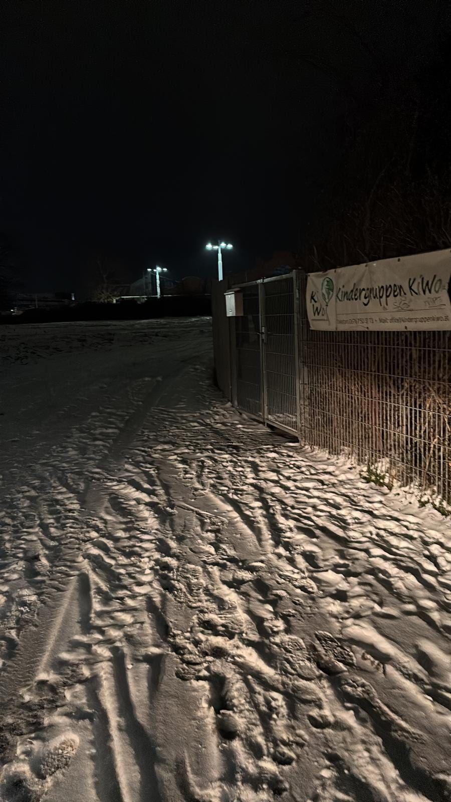 Verschneiter Weg mit Fuß- und Reifenspuren vor einem Gittertor bei Nacht.