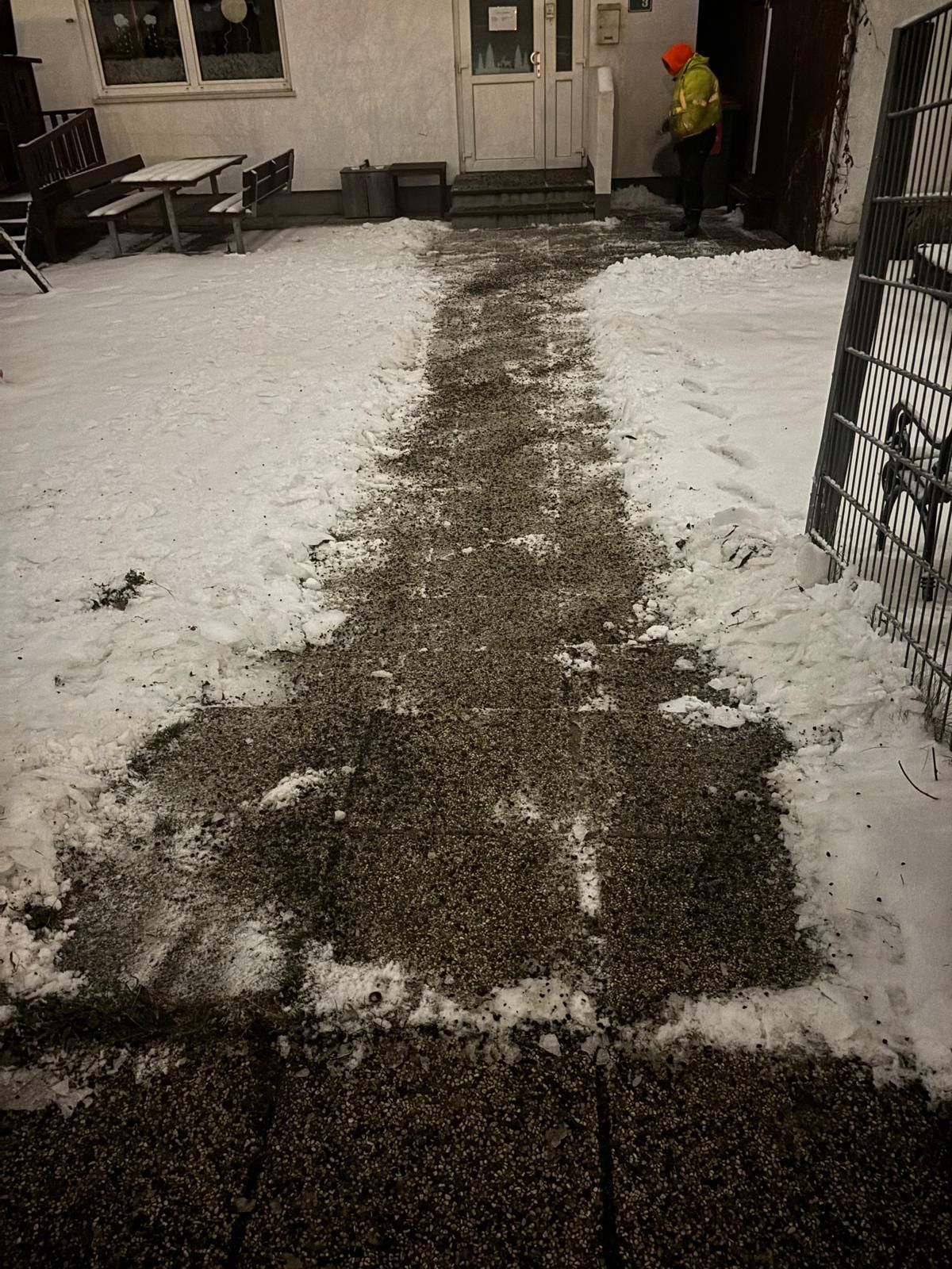 Geräumter und gestreuter Weg durch den Schnee zu einem Hauseingang, im Hintergrund streut ein Mitarbeiter in Warnkleidung.